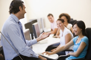 Male instructor speaking to students in computer classroom with smiling students.
